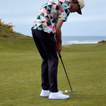 Golfer putting a golf ball with his putter while standing on an oceanside green. He is wearing white, hybrid golf shoes designed by FHADE. 